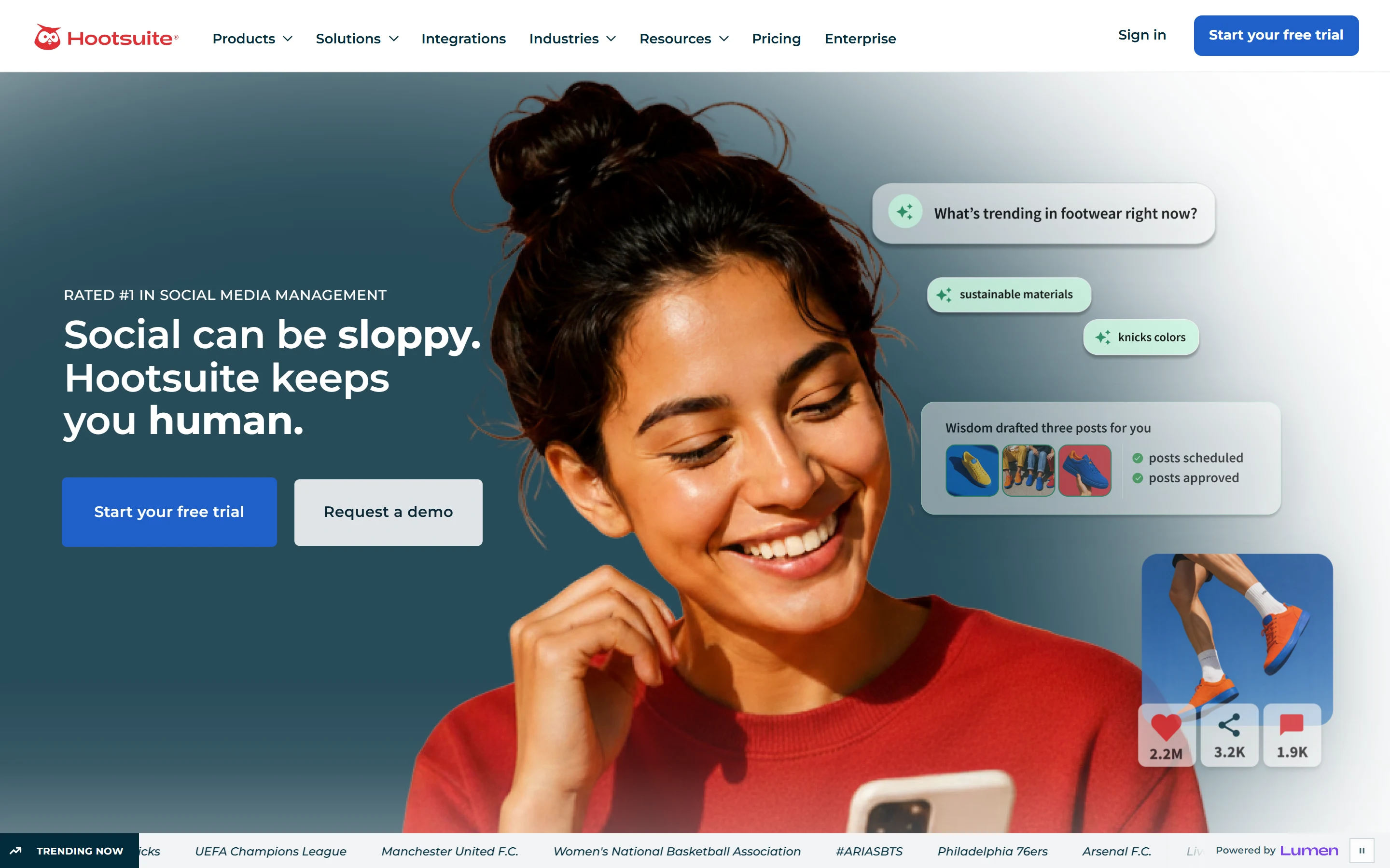Hootsuite homepage hero: the navigation bar with Products, Solutions, Integrations, Industries, Resources, Pricing, Enterprise, Sign in and Start your free trial, the eyebrow “RATED #1 IN SOCIAL MEDIA MANAGEMENT” above the headline “Social can be sloppy. Hootsuite keeps you human.”, Start your free trial and Request a demo buttons, a smiling woman holding a phone, floating cards reading “What’s trending in footwear right now?” and “Wisdom drafted three posts for you” with posts scheduled and posts approved, and a TRENDING NOW ticker marked Powered by Lumen along the bottom